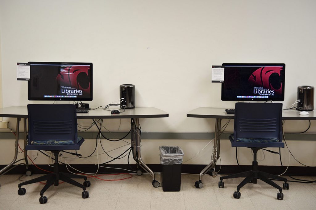Two large monitors of the computers in the Dimensions Lab sit on desks with chairs in front.